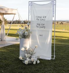 a white sign sitting on top of a lush green field next to a vase filled with flowers