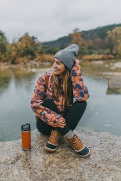 a woman sitting on top of a rock next to a lake wearing a beanie