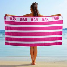 a woman is standing on the beach holding a pink and white towel that says jean jean jean