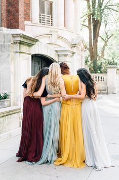 four women in long dresses standing together on the sidewalk with their arms around each other