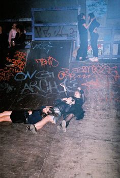 two people laying on the ground in front of a wall with graffiti written all over it