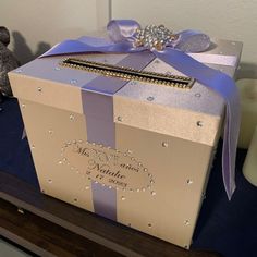 a wedding gift box with a purple ribbon and diamond decoration on the top, sitting on a table next to a teddy bear