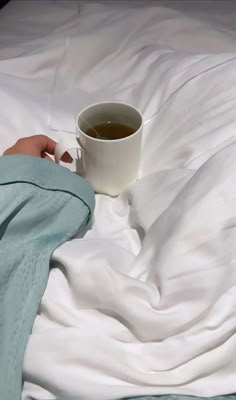 a person's hand holding a cup of tea on top of a white bed