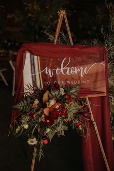 a welcome sign on top of a wooden easel with flowers and greenery around it