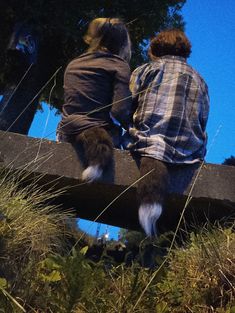 two people sitting on a bench with their feet in the grass and one person standing up