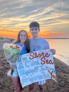 two people standing on the beach holding flowers and a sign