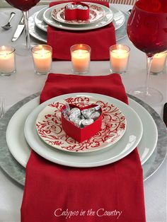 a table set for valentine's day with red napkins and white plates, silverware and candles