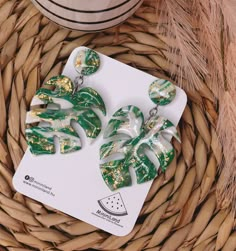 a pair of green and gold leaf earrings sitting on top of a basket next to a cup