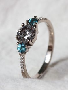 Dainty white gold trilogy ring featuring a central round salt and pepper diamond with teal coloured round side stones on a fine tapering band with little white diamonds set along each side. Grey Gemstones, Gray Gemstones, Black Diamond Ring, Salt And Pepper Diamond