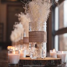 some white flowers are in a glass jar on a wooden table with candles around it