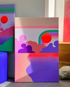 three different colored paintings sitting next to each other on top of a wooden floor near rocks