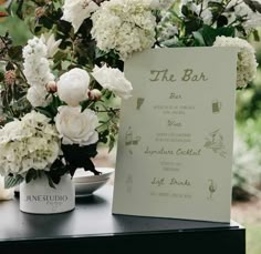 white flowers are in vases next to a menu on a table with wine glasses