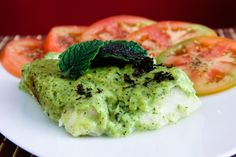 a white plate topped with meat covered in pesto next to tomatoes and cucumbers