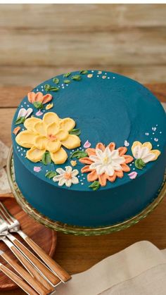 a blue cake with flowers on it sitting on a wooden table next to silverware