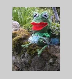 a crocheted frog sitting on top of a rock next to plants and grass