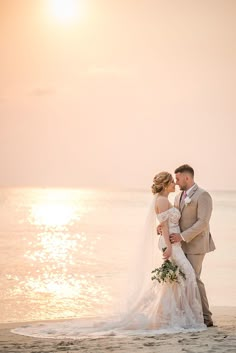 a bride and groom standing on the beach at sunset