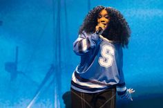 a woman with an afro is singing into a microphone while wearing a blue jersey and black pants