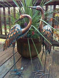 two metal birds are sitting in a potted plant on a wooden deck with palm leaves