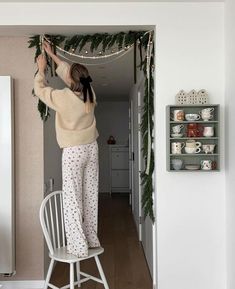 a woman hanging garland on the wall in her kitchen while wearing pajamas and holding onto a chair