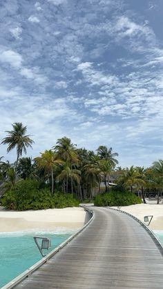 a wooden bridge over water leading to a sandy beach with palm trees in the background