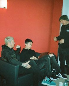 two men sitting on couches in front of a red wall talking to each other