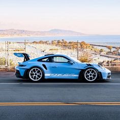 a blue sports car parked in front of a fence with the ocean and mountains in the background