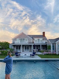 a woman standing in front of a swimming pool next to a large white house with an open porch