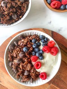 granola with berries and chocolate chips in a bowl on a wooden cutting board next to two bowls of yogurt