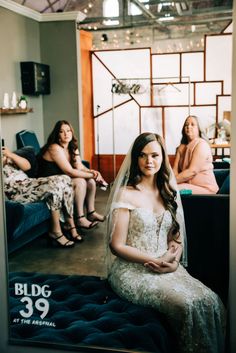 a woman in a wedding dress is sitting on a bed with other women behind her