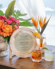 an arrangement of flowers and drinks on a wooden table next to a vase filled with oranges