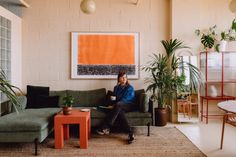 a woman sitting on top of a green couch in a living room next to potted plants