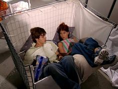 two people laying on a bed in a room with white walls and an open laundry basket