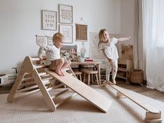 two children playing on a wooden slide in a room with white walls and flooring