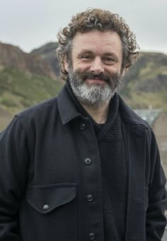 a man with curly hair and beard standing in front of some mountains wearing a black jacket