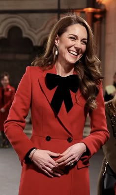 a woman wearing a red coat and black bow tie smiles as she walks down the runway