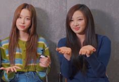 two young women sitting next to each other in front of a wall with one pointing at the camera