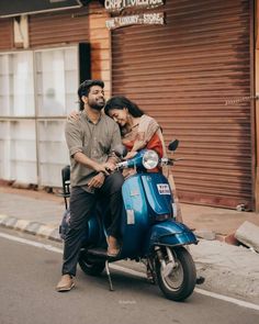 a man and woman riding on the back of a blue scooter down a street