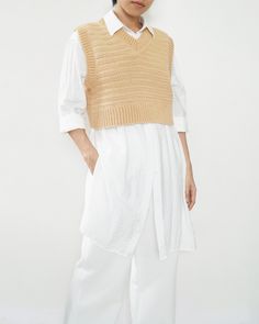 a woman standing in front of a white wall wearing a tan sweater and white pants