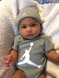 a baby laying on top of a bed wearing a gray shirt and hat with a white basketball logo