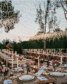 an outdoor dinner table set up with place settings and lite - up trees in the background