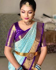 a woman in a purple and blue saree with gold jewelry on her neck, standing next to a couch