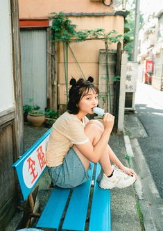 a woman is sitting on a blue bench drinking from a water bottle in an alleyway