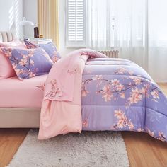 a bed covered in pink and purple comforter next to a window with white curtains