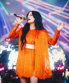 a woman in an orange dress holding a microphone and singing into the air at a concert