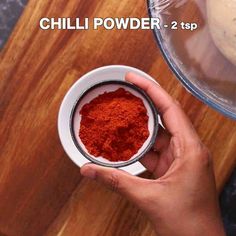 a person holding a bowl filled with red powder on top of a wooden table next to a blender