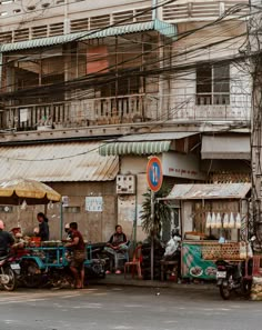 some people are sitting at an outdoor food stand on the side of the road in front of a building with scaffolding