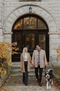 a man and woman holding hands while walking their dog in front of an old building