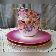 two tea cups and saucers with flowers on them sitting on a lace tablecloth