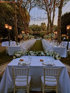 a long table set up with white linens and place settings for an outdoor dinner