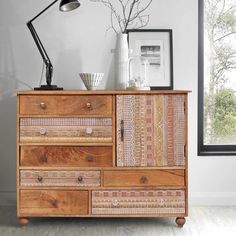 a wooden dresser with drawers in front of a window next to a lamp and pictures on the wall